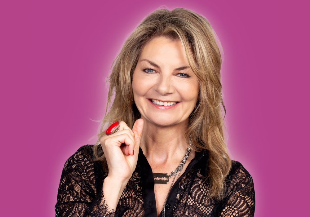 Comedian Jo Caulfield stands in front of a dark pink background, she is smiling at the camera and has one hand up next to her face.