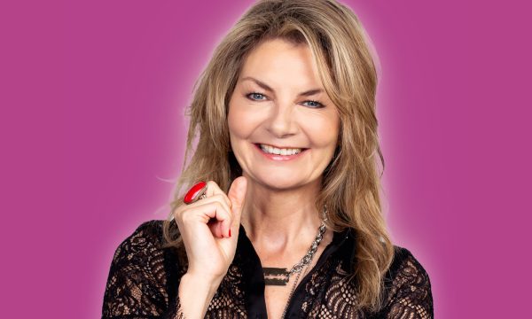 Comedian Jo Caulfield stands in front of a dark pink background, she is smiling at the camera and has one hand up next to her face.