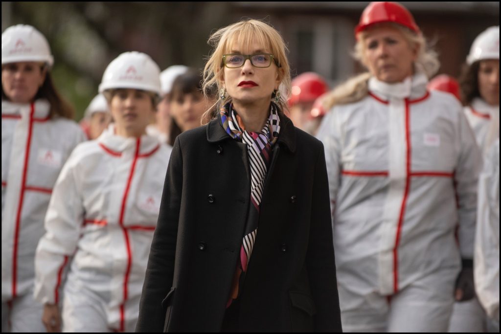 A blonde-haired woman with glasses, wearing a smart, dark coat, walks ahead of a group all wearing protective clothing and hard hats