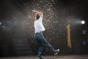 Luca, wearing blue jeans and a white t-shirt, dances as confetti falls around her. A yellow dress hangs from a coathanger in the background.