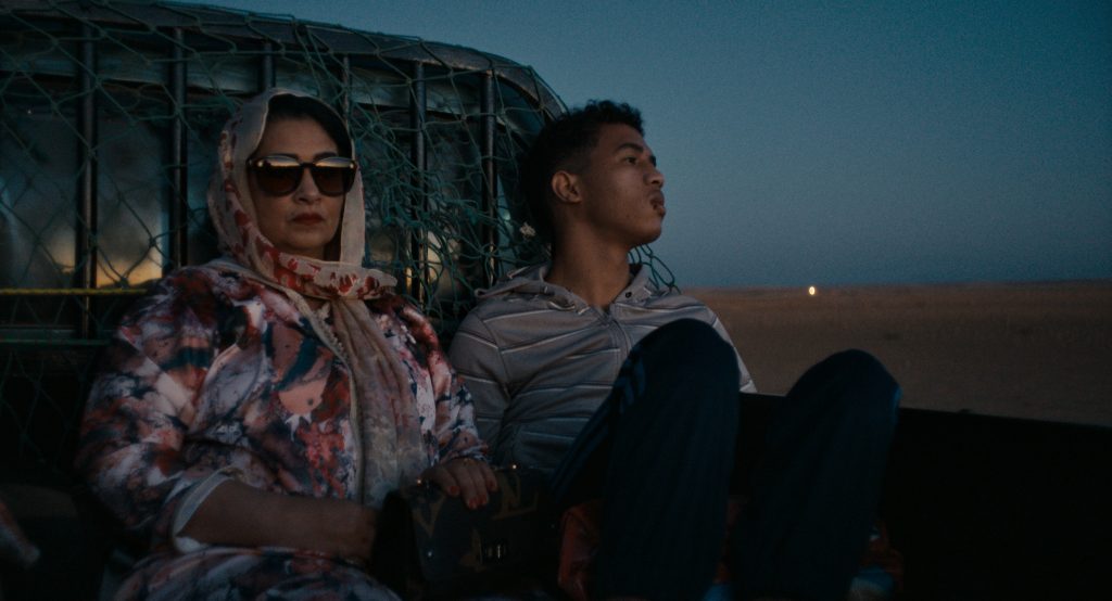 A mother and son sit in the back of a pick-up truck in the middle of a dessert. The son is looking into the distance, he has a cut lip. The mother is looking forward, her face is obscured by large sunglasses, she has a floral scarf wrapped loosely over her head and is carrying a designer handbag.