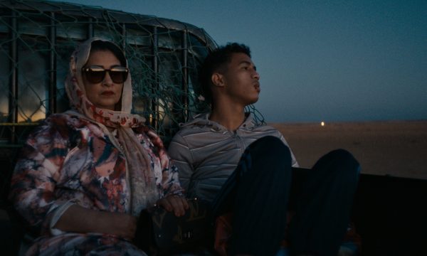 A mother and son sit in the back of a pick-up truck in the middle of a dessert. The son is looking into the distance, he has a cut lip. The mother is looking forward, her face is obscured by large sunglasses, she has a floral scarf wrapped loosely over her head and is carrying a designer handbag.