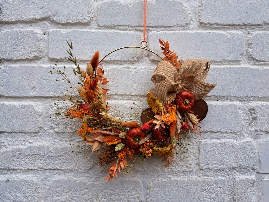 A dried flower autumn wreath, full of orange and red dried foliage and flowers set around a gold hoop, hangs in front of white painted brick wall.