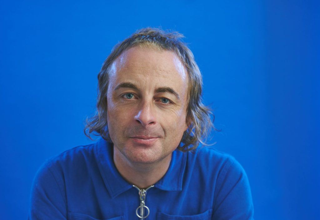 A portrait of comedian Paul Foot, a man with mid-length blonde hair wearing a bright blue shirt. He is looking directly into the camera and smiling. The background is also bright blue.