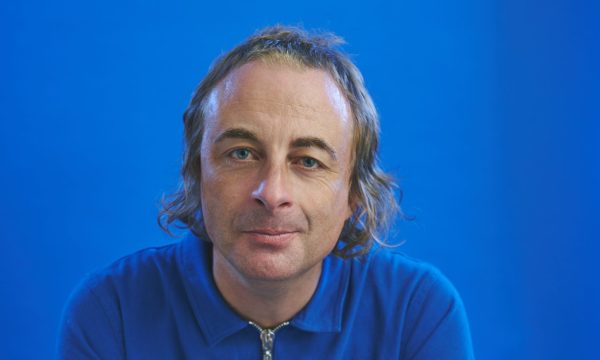 A portrait of comedian Paul Foot, a man with mid-length blonde hair wearing a bright blue shirt. He is looking directly into the camera and smiling. The background is also bright blue.