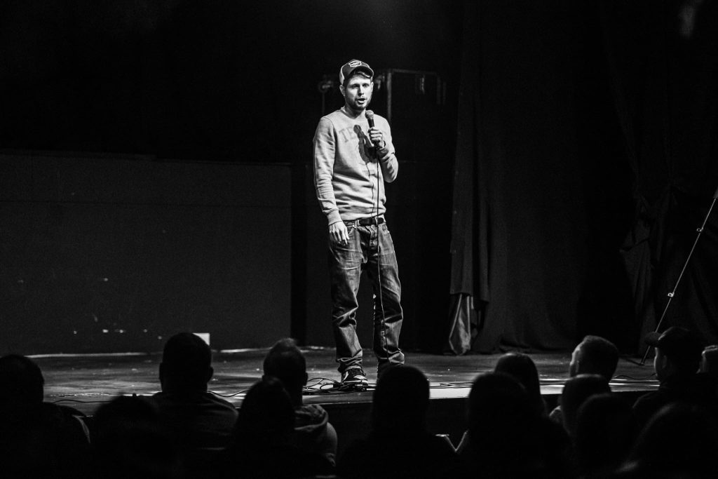 A black and white photo of comedian Phil Chapman, he is standing at the front of the stage holding a mic. The front row of the audience is visible at the bottom of the image.
