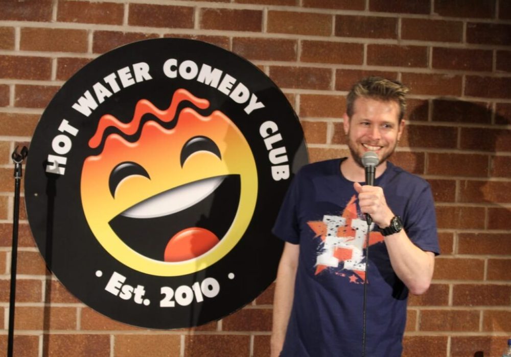 Comedian Phil Chapman, a white man with short brown hair, is holding a microphone and standing in front of the Hot Water Comedy Club logo. He is grinning broadly.