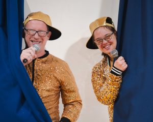 Gav and Megan stand with gold jackets and hats on popping out of curtains