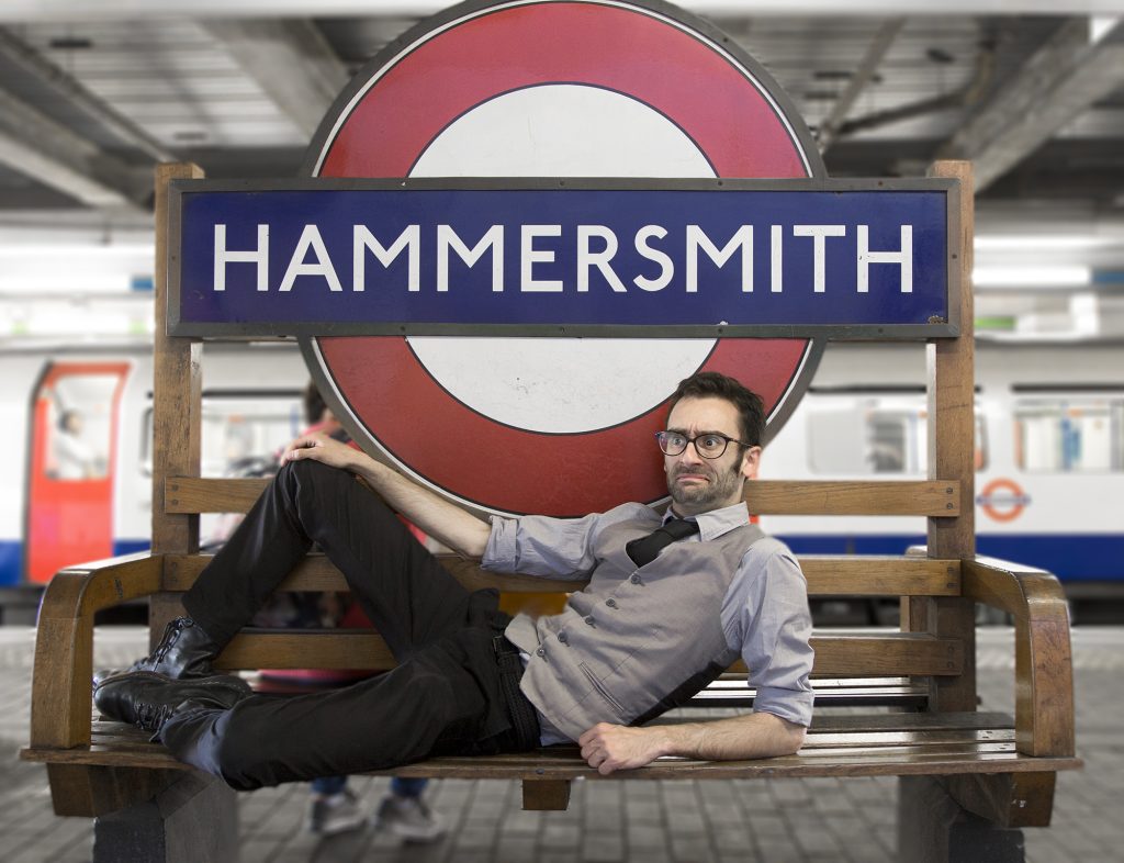 Comedian MC Hammersmith reclining on a bench in front of a tube sign reading Hammersmith. He is wearing a waistcoat, tie and trousers.