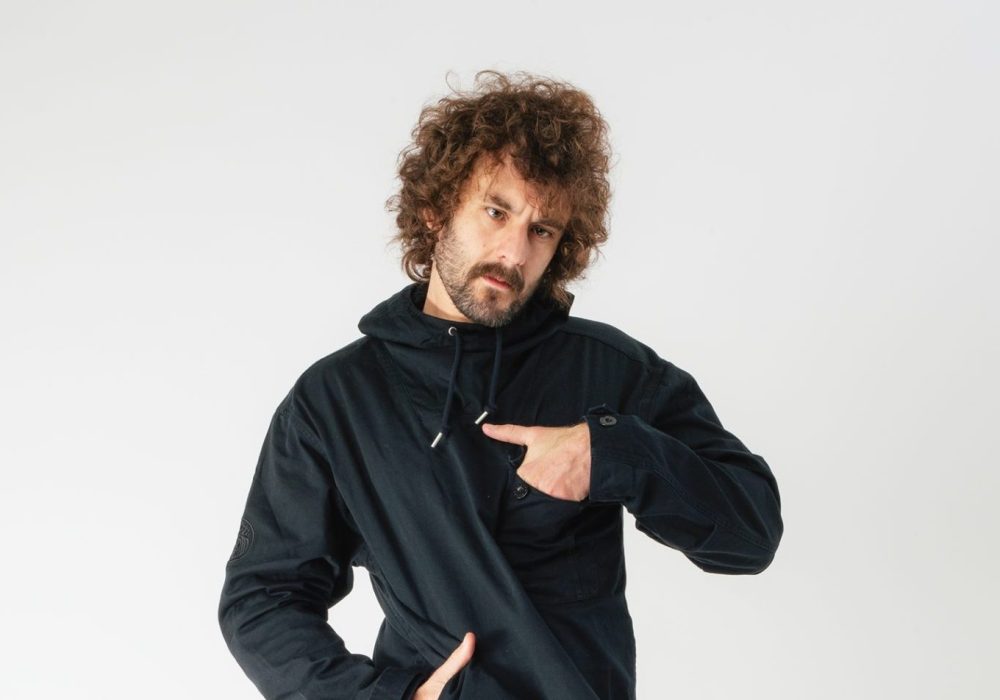 Comedian Josh Pugh, he is standing in front of a white background with one hand in his pocket on his waist, and the other hand in a pocket on his chest.
