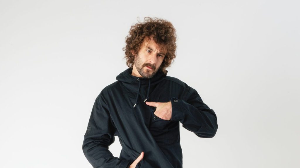 Comedian Josh Pugh, he is standing in front of a white background with one hand in his pocket on his waist, and the other hand in a pocket on his chest.