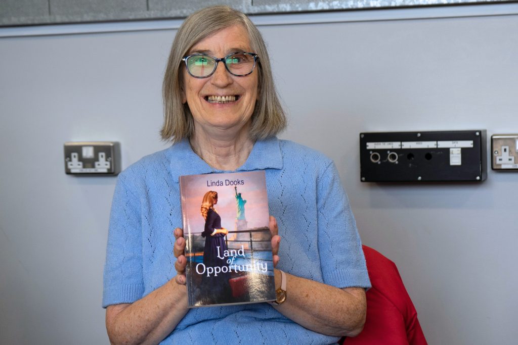 Creative Writing participant and published author Linda Dooks. Linda is a white woman, with shoulder length blonde-grey hair. She is holding a copy of her newly published book, and is smiling proudly.