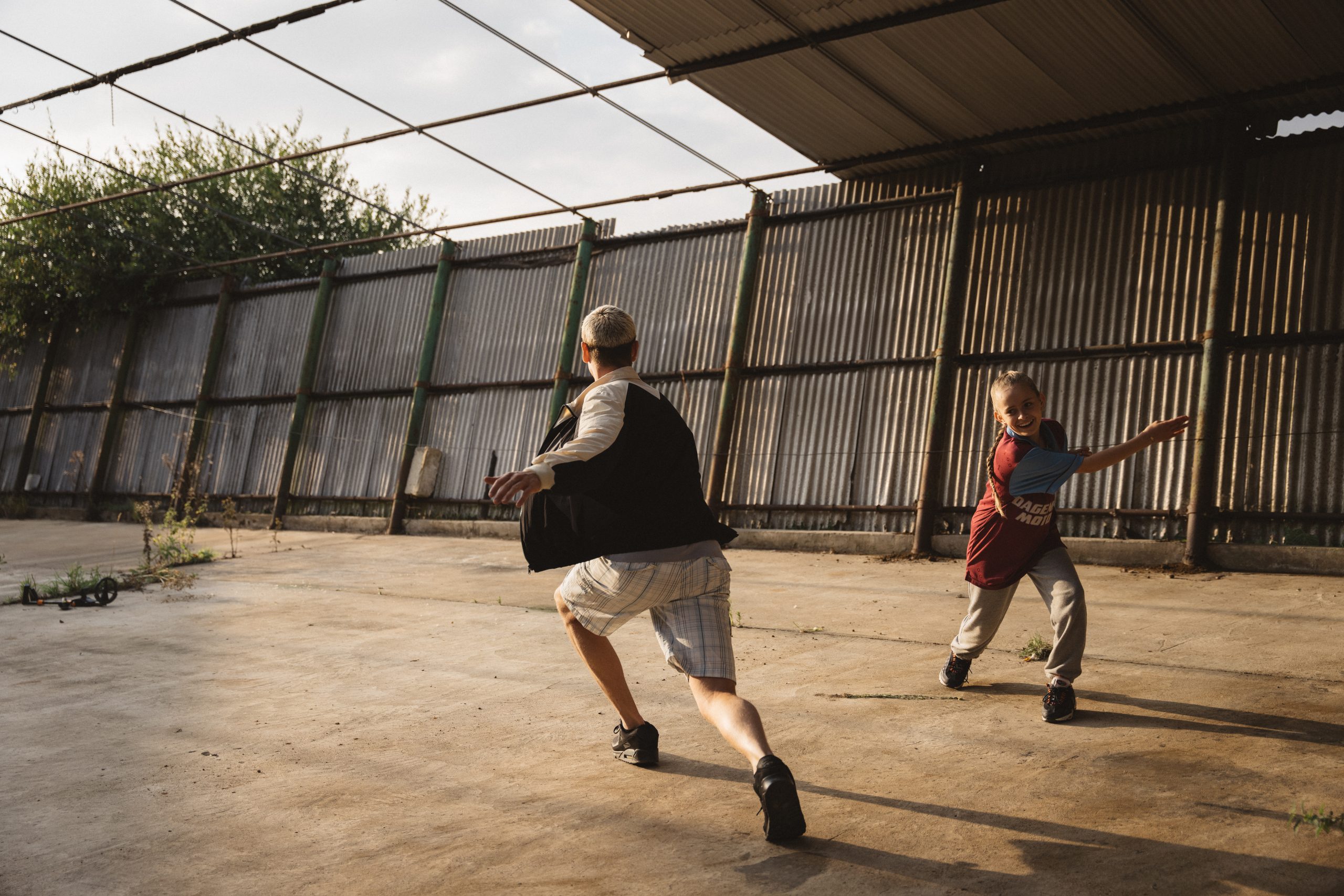 Image of a rather and daughter running around playfully in an area with a concrete floor, and corrugated metal walls, all bathed in a warm light.