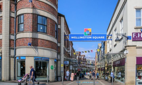 A shot looking down the lane of outdoor shopping centre, Wellington Square, in Stockton. It's a sunny day with a bright blue sky, and there is bunting strung between the buildings on the two sides of the lane. Plenty of people are out shopping.