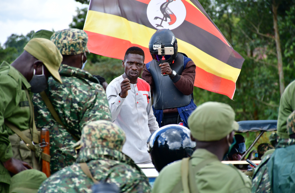 Image of Bobi Wine a black Ugandan Man in a white shirt with a red shoulder, he is speaking to a group of men in camouflage. Over his shoulder is someone in a protective jacket and helmet that says press holding a camera up to film.