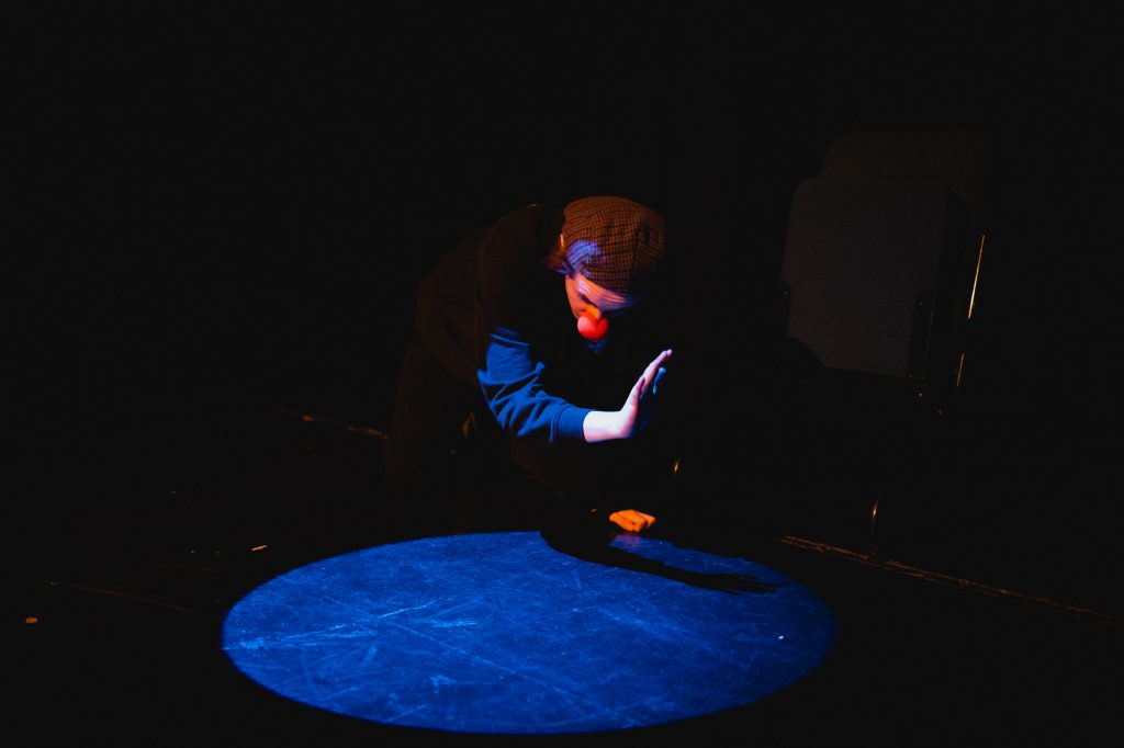 A performer is kneeling, and is half in / half out of a spotlight, cretaing a shadow with their hand in the light. They are wearing a checked flat cap (back to front), and a clown's red nose