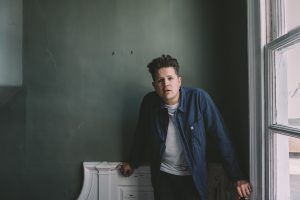 Luke, wearing a blue shirt, white t-shirt and dark trousers, stands between a window and a fireplace, with one hand leaning on each.