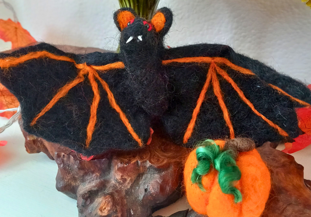A black needle felt bat, with orange detailing, and a small orange pumpkin, with green curly tendrils.