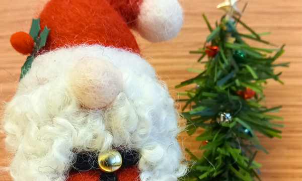 A needle felt Santa Gnome, next to a tree made of tinsel. The gnome has a white beard, and only has his nose visible. He's wearing a santa outfit.