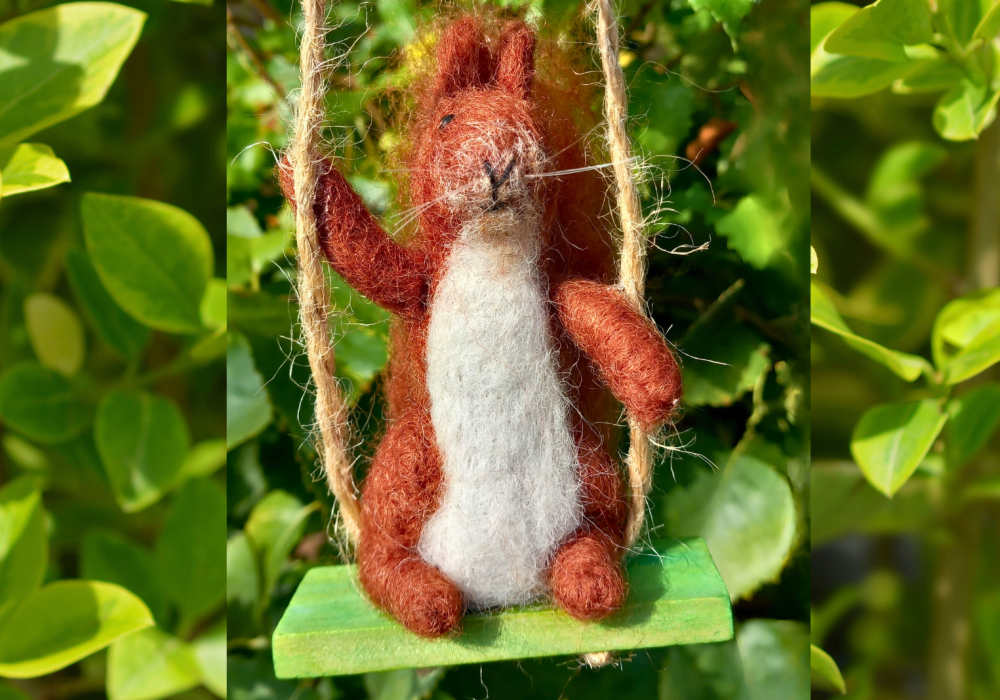 A needle felted red squirrel, sitting on a swing in front of a green bush.