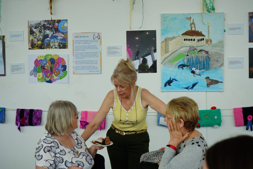 Image at an exhibition showing a range of artwork created at Staying Out. The gallery space has white walls and incudes a range of artwork including collage, writing, and textiles. In front of the exhibition three women, two sitting, and one standing, are engaged in a conversation together.