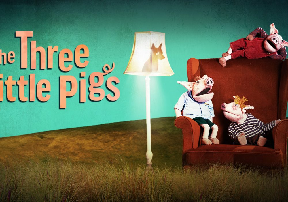 On the left hand side of the image is the show title, 'The Three Little Pigs'. On the right hand side are three puppet-style pigs, wearing clothes, sitting in various spots on the same large, red armchair. A standard lamp, switched on, is to the side of the chair. The shadow of a wolf is looking down at the pigs from inside the lampshade.