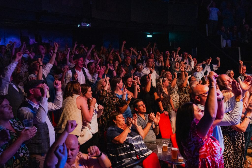 Image shows a large crowd of people facing out of shot towards a strong light, they are all singing, dancing, and smiling.
