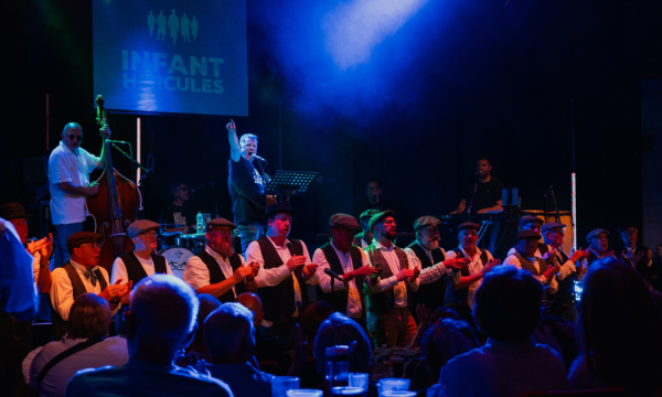 Photo of a gig at ARC Stockton. Mike McGrother points from the stage out into the audience, he is singing into a microphone. Behind him are The Wildcats of Kilkenny, in front Infant Hercules men's choir lines the front of the stage.