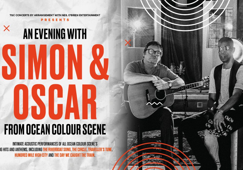 Simon & Oscar sit side by side, Simon holds an acoustic guitar, Simon sits on a cajon holding a tambourine