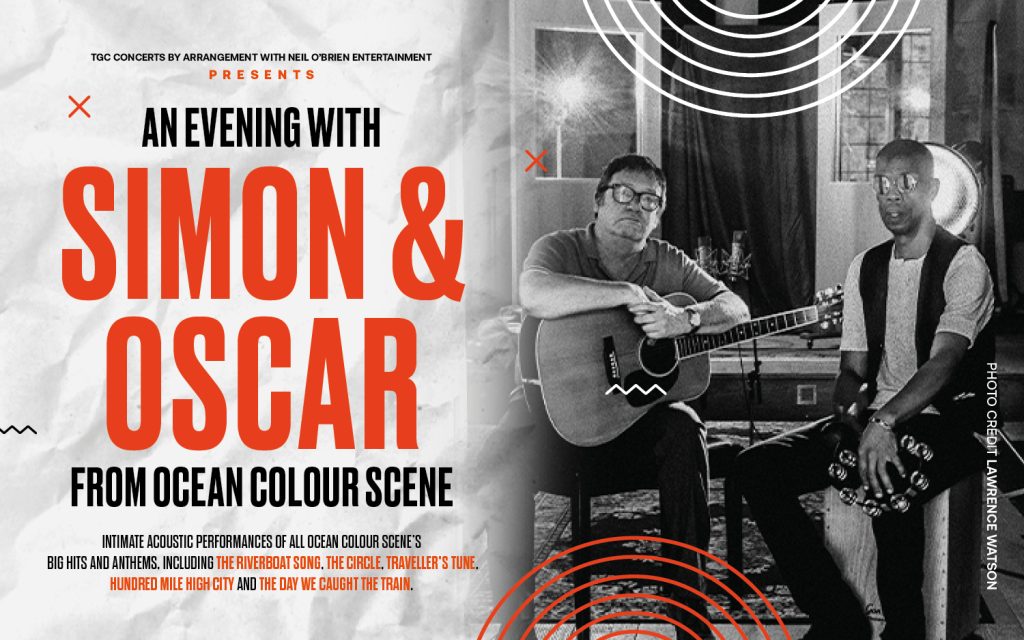 Simon & Oscar sit side by side, Simon holds an acoustic guitar, Simon sits on a cajon holding a tambourine