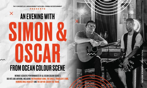 Simon & Oscar sit side by side, Simon holds an acoustic guitar, Simon sits on a cajon holding a tambourine