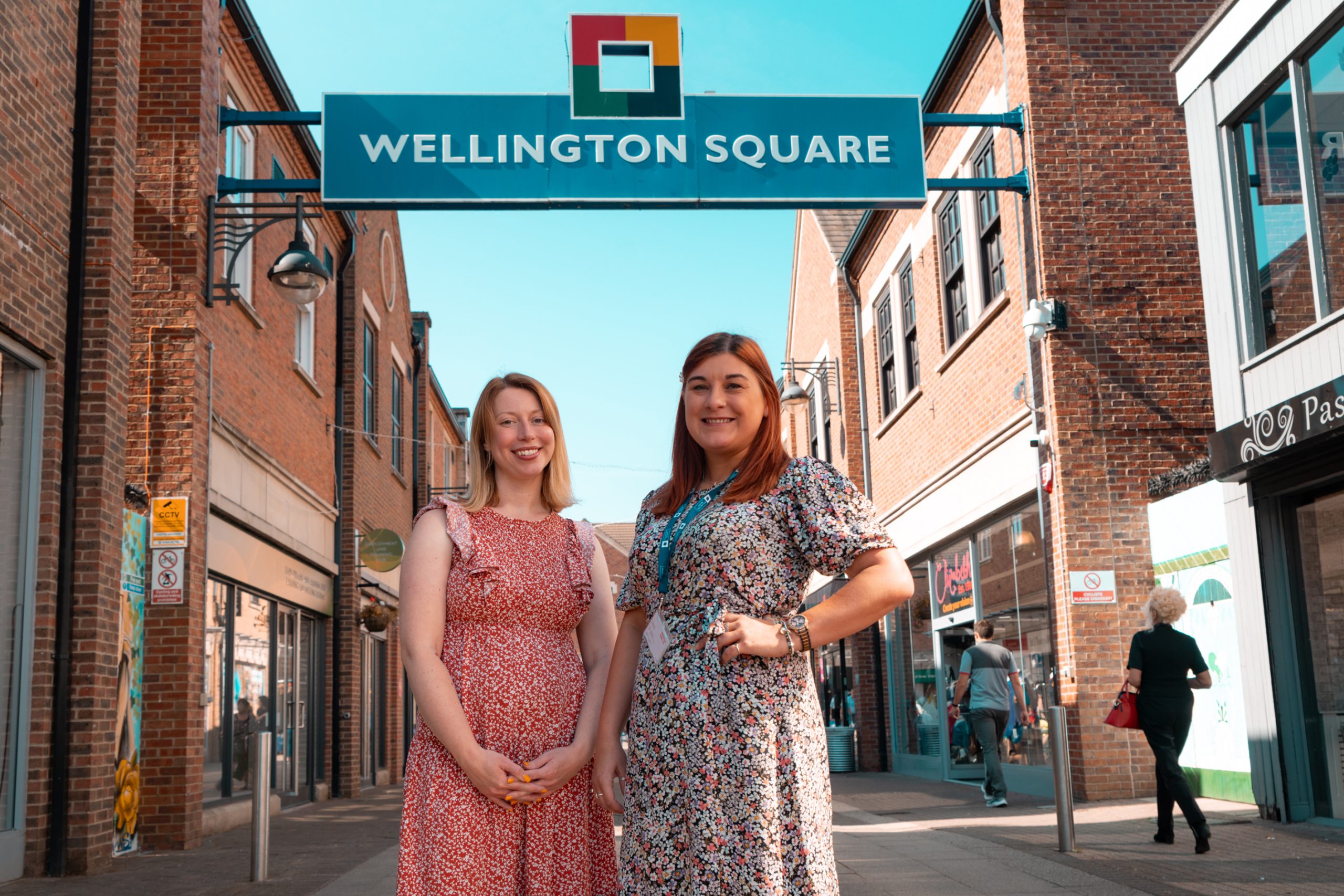 Image shows two women looking to the camera in a shopping centre with rows of shops on either side. Above them is a logo of a square made from four brightly coloured right angles, and the words Wellington Square.