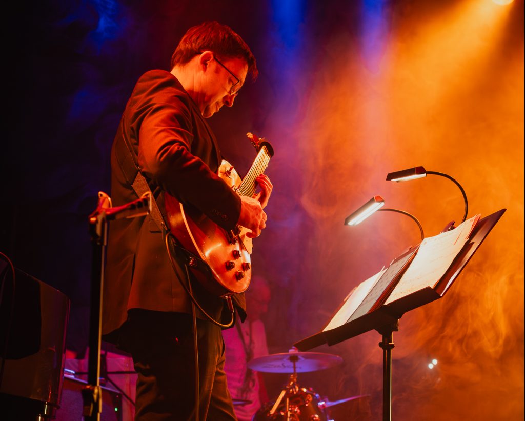 Paul Donnelly stands playing guitar in front of a music stand with band mates playing behind on stage including bass guitar, sax and drums