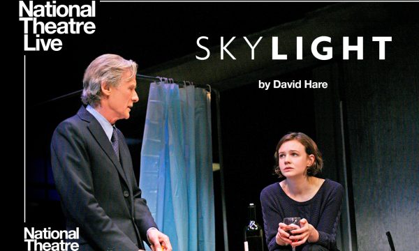 man in a suit stood facing a woman sat with glass of red wine National Theatre Live Skylight