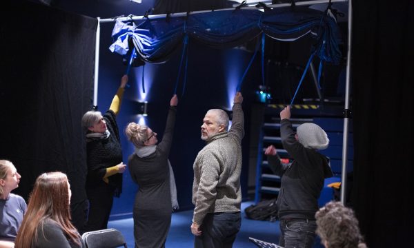 Four people are standing and reaching up to release a blue piece of material tied to a metal bar above them. A small, seated audience watches on.