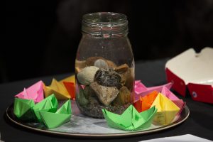 A large jar, filed with water and half-filled with stones, stands in the middle of a plate. Water from the jar has overflowed onto the plate, and brightly coloured, folded paper boats are floating on the plate.