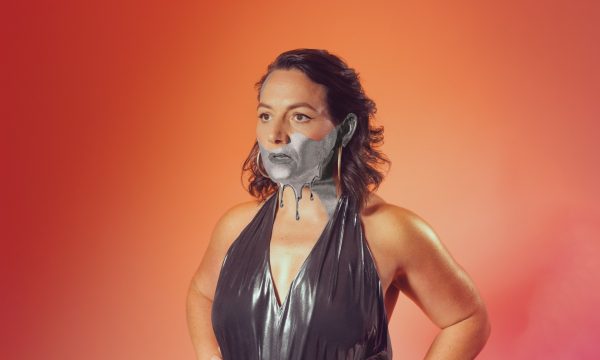 A half body shot of comedian Jessica Fostekew. She stands against an ombre orange and pink background, wearing a dark grey metallic dress. The bottom half of her face is black and white.