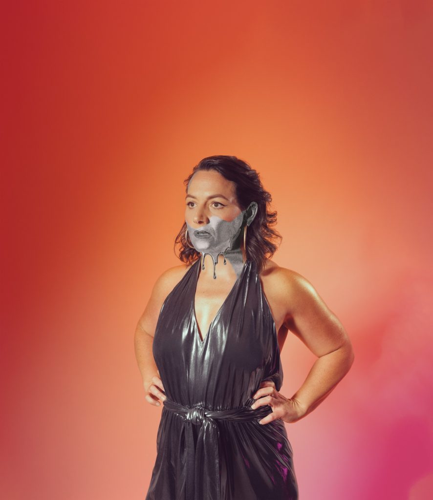 A half body shot of comedian Jessica Fostekew. She stands against an ombre orange and pink background, wearing a dark grey metallic dress. The bottom half of her face is black and white.
