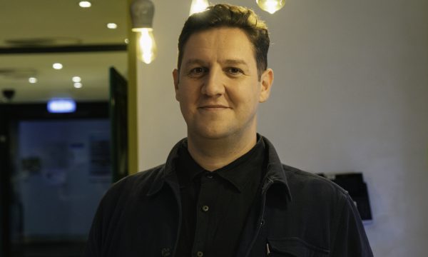Image of Alexander Ferris, a white man with dark hair he is standing in the foyer of ARC Stockton Arts Centre, and is smiling looking directly towards the camera.