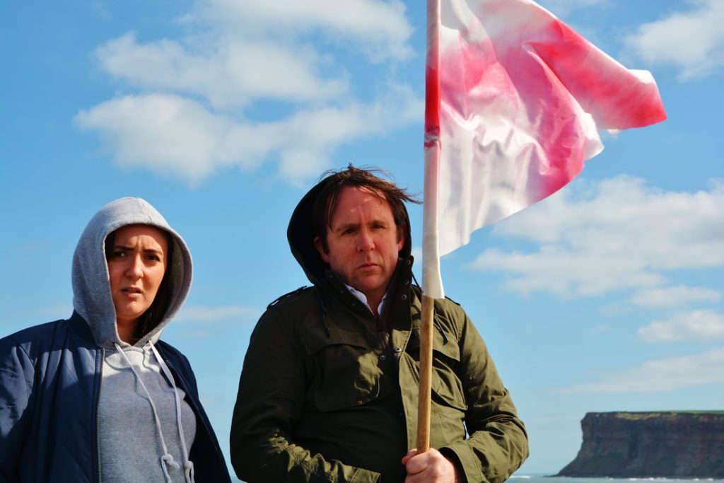 A woman and a man (holding an England flag on a pole) with sea and cliffs in the background