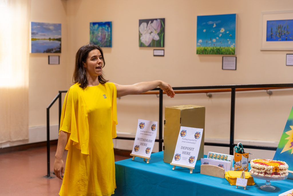 Alice Roots, wearing a bright yellow dress, is standing in front of a table with signs saying 'withdraw here' and 'deposit here' and is speaking to people off camera.