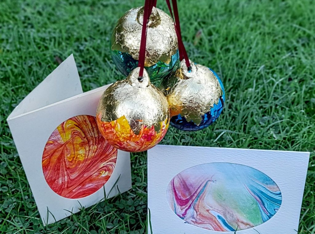Three brightly coloured marbled baubles with gold foil designs are hanging above two marbled paper cards.