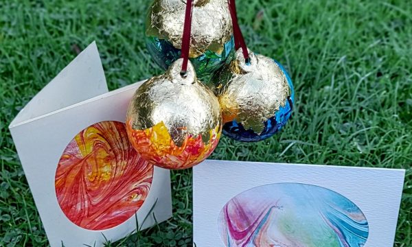Three brightly coloured marbled baubles with gold foil designs are hanging above two marbled paper cards.