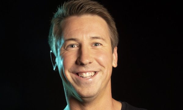 A portrait of comedian Carl Hutchinson, he is in front of a black background and he is smiling broadly.