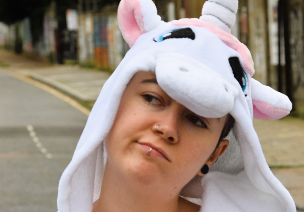 Headshot of Jules the Poet out in the street. Jules is looking away from the camera, and is wearing a unicorn outfit with the hood up.