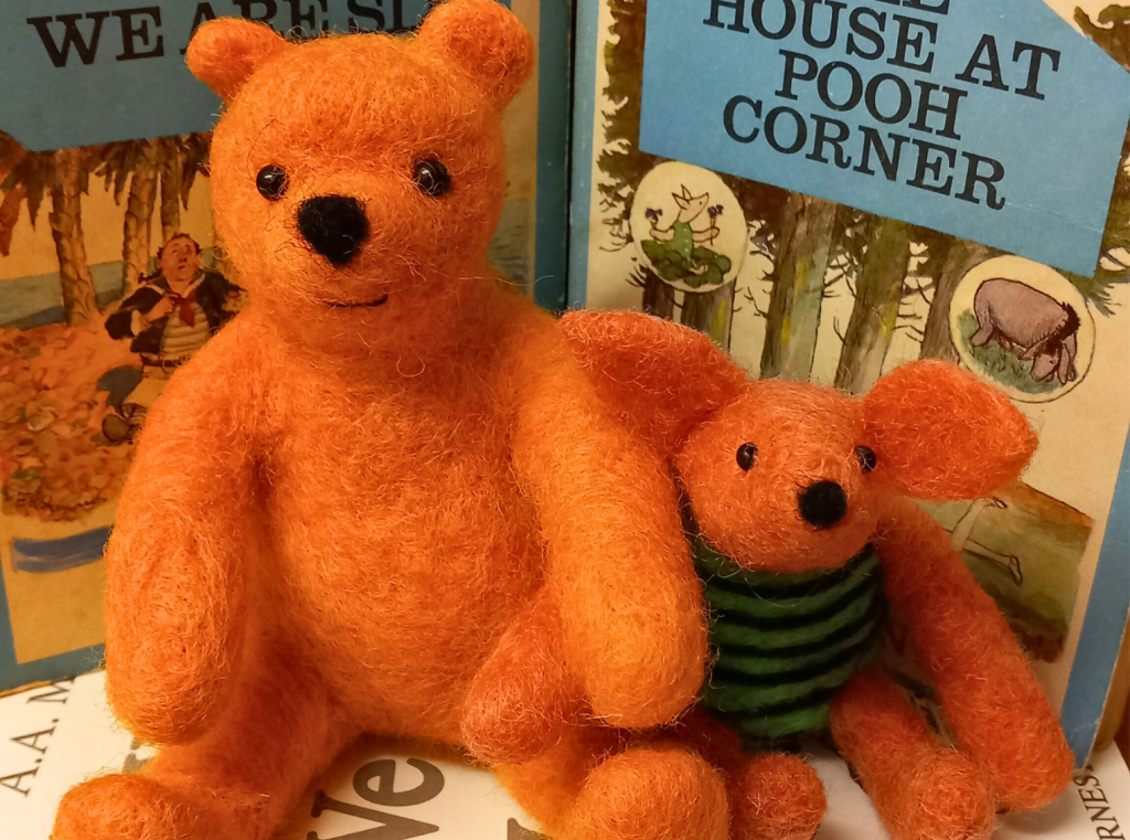 A yellow needle felt bear sits beside a pink needle felt piglet, they are both in front of the cover of AA Milne's House at Pooh Corner