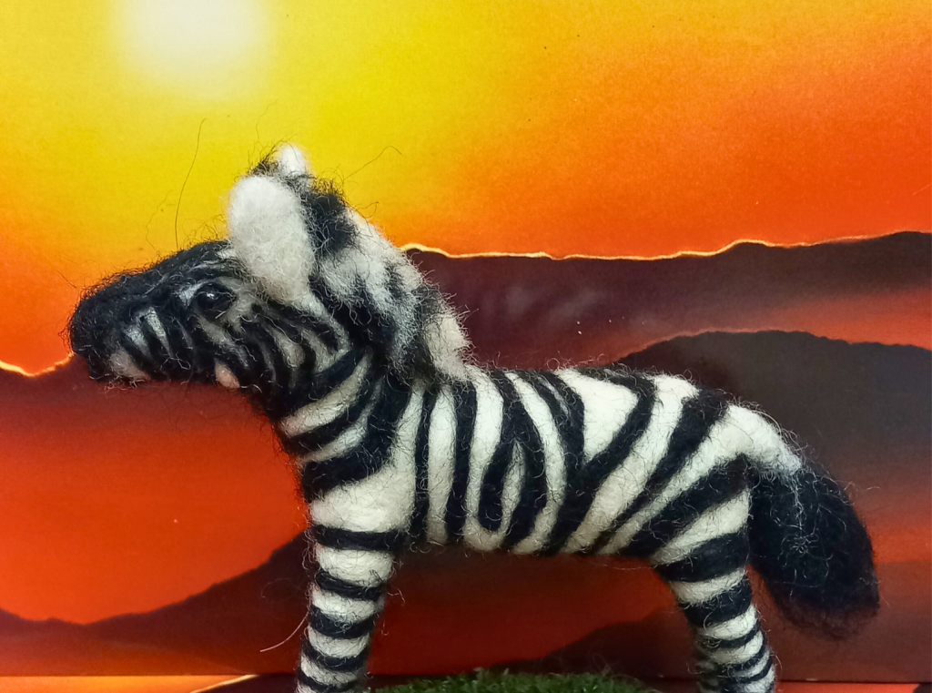 A felted zebra stands in front of a painting of a desert at sunset.