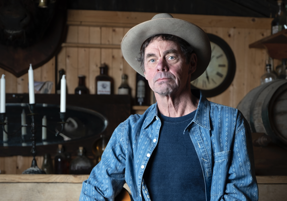 Comedian Rich Hall stands in a wooden bar, he is wearing a grey suede cowboy hat and holding an acoustic guitar.