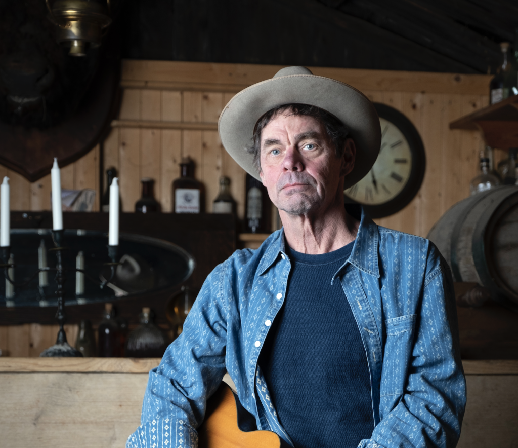 Comedian Rich Hall stands in a wooden bar, he is wearing a grey suede cowboy hat and holding an acoustic guitar.