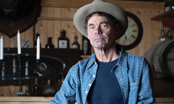 Comedian Rich Hall stands in a wooden bar, he is wearing a grey suede cowboy hat and holding an acoustic guitar.